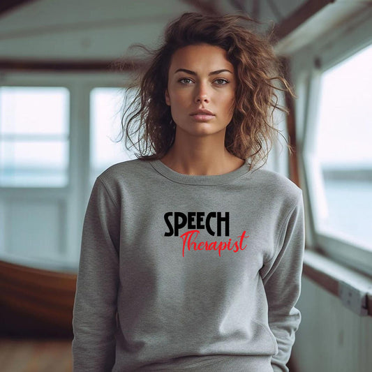 Person wearing a grey Speech Therapist statement clothing crew neck sweatshirt with bold black and red text, standing in a modern interior setting. Premium DTG print quality, fast delivery, and free worldwide shipping by Yebber.