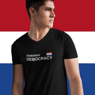 Person wearing a black Remember Democracy Netherlands statement clothing T-shirt with white text and a small Dutch flag, standing in front of a large Dutch flag background.