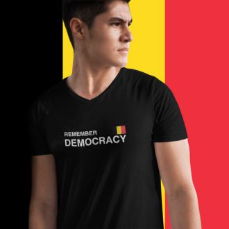 Person wearing a black Remember Democracy Belgium statement clothing T-shirt with white text and a small Belgian flag, standing in front of a large Belgian flag background. 