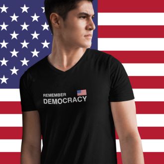 Person wearing a black Remember Democracy USA statement clothing T-shirt with white text and a small USA flag, standing in front of an American flag background. 