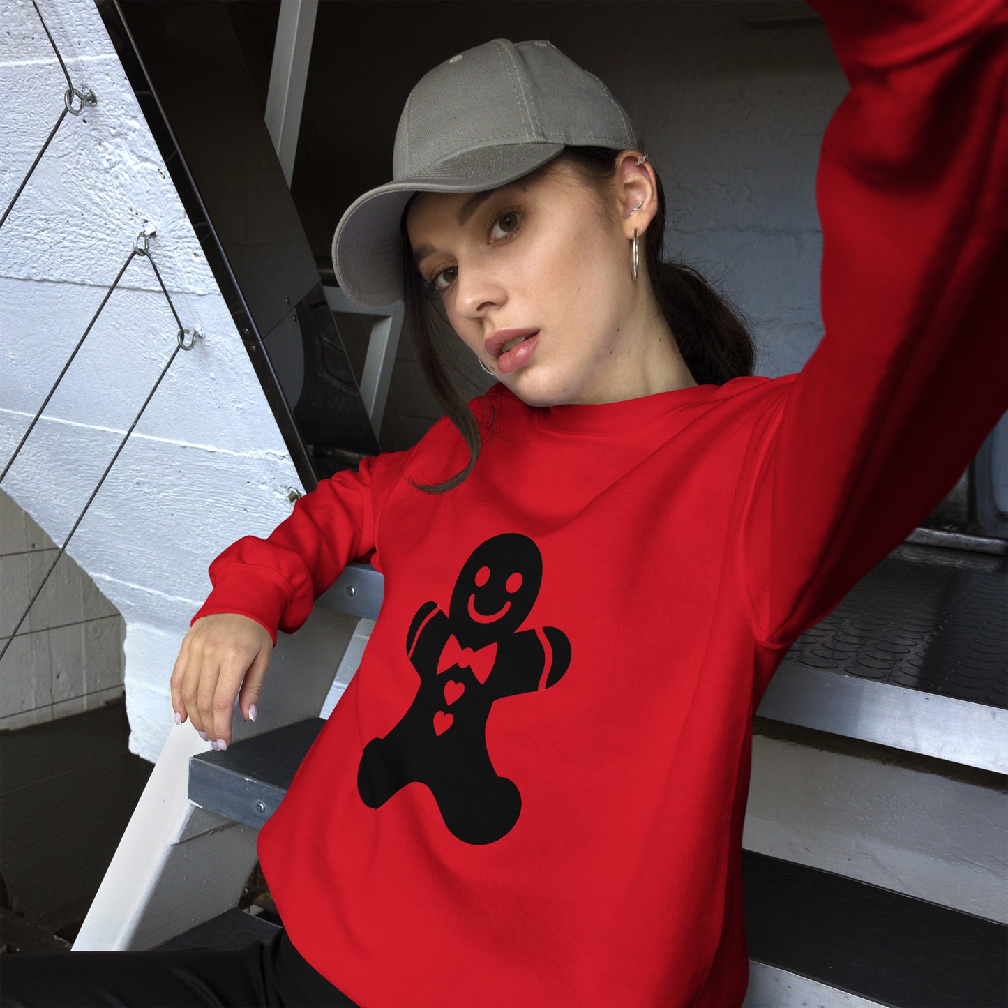 Person wearing a red Minimalistic Christmas statement clothing sweatshirt with a black gingerbread man design, making a playful peace sign gesture while sitting at a stairway.